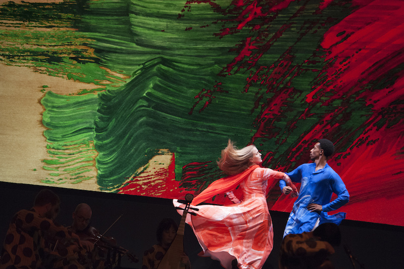 A man in a blue tunic swings a woman in a coral dress by the arm. Her blond hair blows behind her.